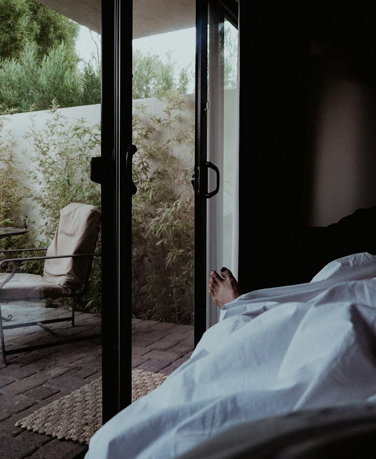 Pessoa descansando na cama, ao lado de uma porta de vidro aberta para o jardim